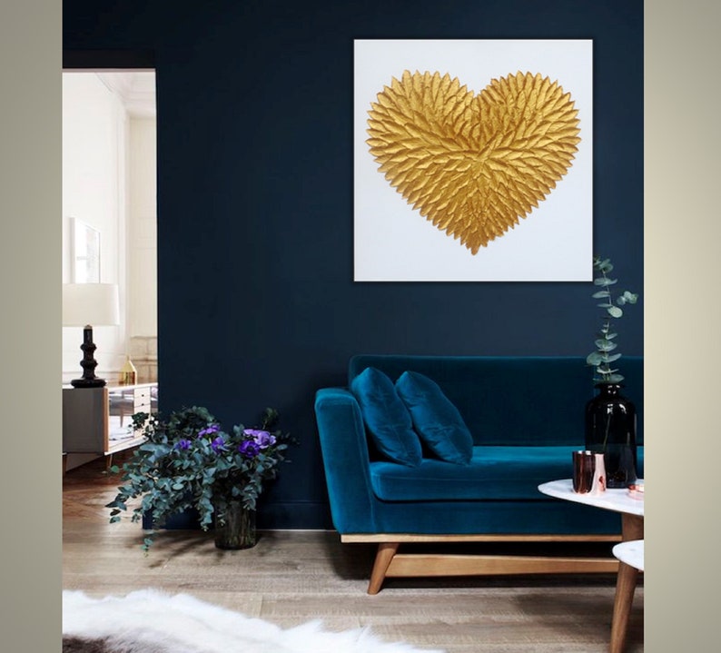 May include: A square wall art piece featuring a textured, three-dimensional gold heart composed of layered leaf-like shapes centered on a plain white background. The artwork is displayed on a dark navy blue wall above a deep blue velvet sofa.