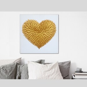 May include: A square wall art piece featuring a textured, three-dimensional golden heart shape centered on a plain white background. The heart is composed of numerous small, overlapping leaf-like elements that create a dense, metallic gold surface.