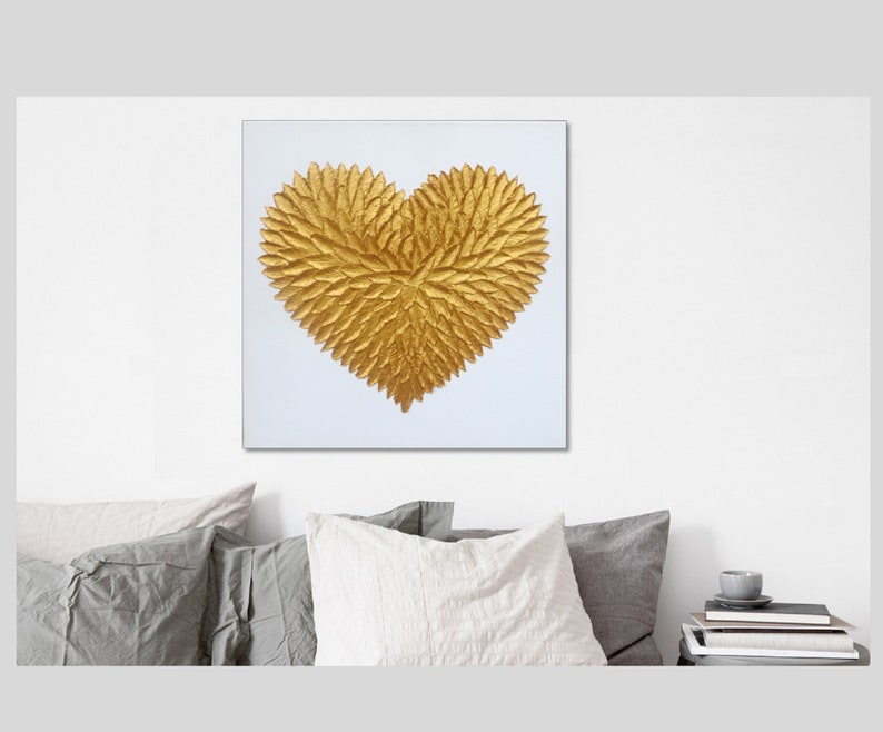 May include: A square wall art piece featuring a textured, three-dimensional golden heart shape centered on a plain white background. The heart is composed of numerous small, overlapping leaf-like elements that create a dense, metallic gold surface.