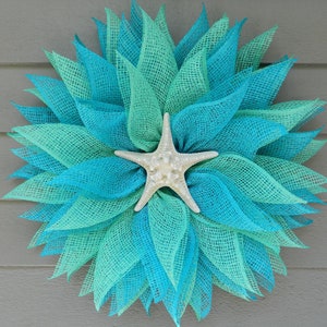 May include: A teal and turquoise mesh wreath with a white starfish in the center. The wreath is made of burlap and has a rustic, coastal feel.