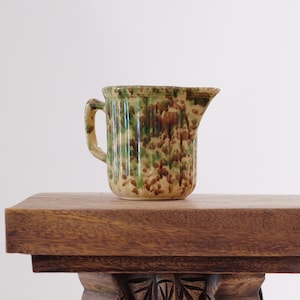 May include: A small, vintage ceramic pitcher with a handle and spout. The pitcher features a speckled pattern of brown and green on a cream-colored base. It is displayed on a wooden surface.