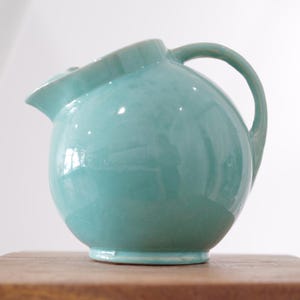 Op de afbeelding: Een turquoise keramische kan met een afgeronde body, een gebogen handvat en een tuit. De kan heeft een glanzende afwerking en staat op een houten oppervlak. Het ontwerp is eenvoudig en elegant, geschikt voor het serveren van dranken.