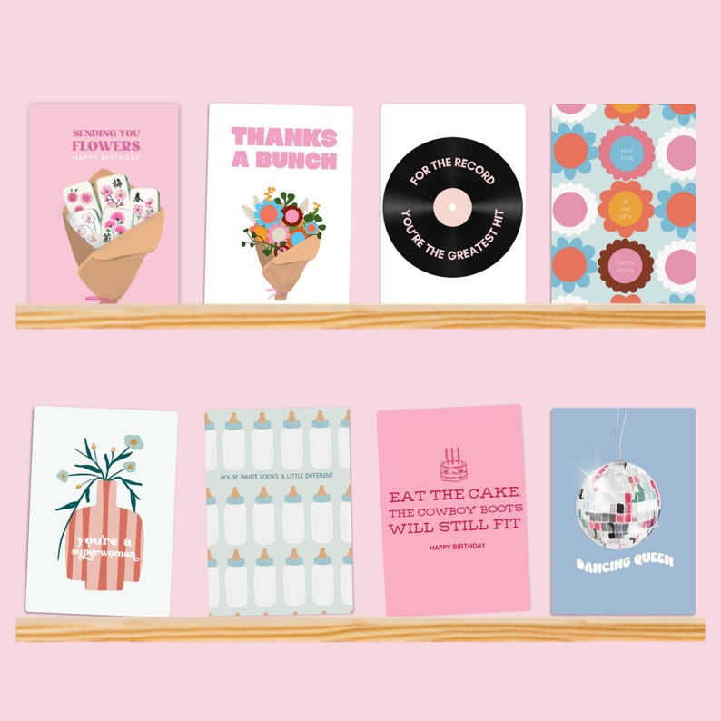 May include: Eight rectangular greeting cards arranged on two wooden shelves. Designs include floral bouquets, a vinyl record with text, colorful flower patterns, a vase, baby bottles, a birthday cake with text, and a disco ball with text.