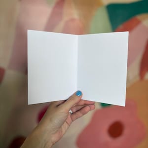 May include: A blank, rectangular, white greeting card held open to reveal two empty, smooth, matte-finish interior panels. The card is constructed from thick, high-quality paper stock, featuring a clean, sharp center fold and crisp, straight edges.