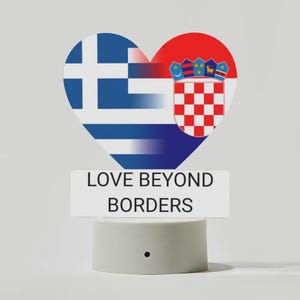Könnte beinhalten: Ein herzförmiges Design mit den Flaggen Griechenlands und Kroatiens, mit dem Text "LOVE BEYOND BORDERS" auf einem weißen Rechteck. Das Design befindet sich auf einer kleinen, weißen, zylindrischen Basis.