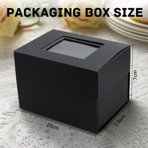 May include: Black cardboard box with a window, measuring 10cm by 7.5cm by 7cm.  The box is ideal for packaging small items.