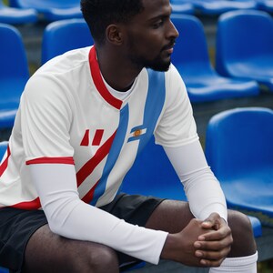 May include: A man wearing a white and blue soccer jersey with red stripes and the flag of Uruguay on the left sleeve. He is sitting on a blue bleacher.