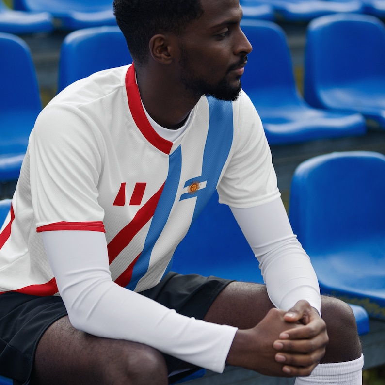 May include: A man wearing a white and blue soccer jersey with red stripes and the flag of Uruguay on the left sleeve. He is sitting on a blue bleacher.