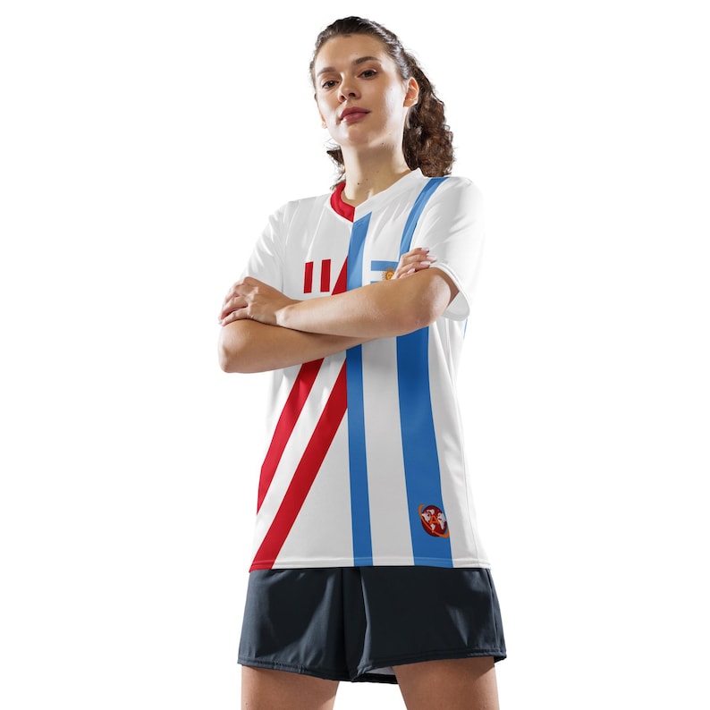 May include: White soccer jersey with red and blue stripes and a small logo on the right side. The jersey has a short sleeve design.