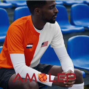May include: A person wearing a white and orange football shirt with the American and Dutch flags on the chest. The shirt has a white long-sleeved undershirt. The person is sitting on a blue stand.