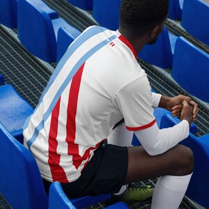 May include: A person wearing a white and red striped jersey with blue accents. The jersey has a white collar and short sleeves. The person is sitting on blue bleachers.