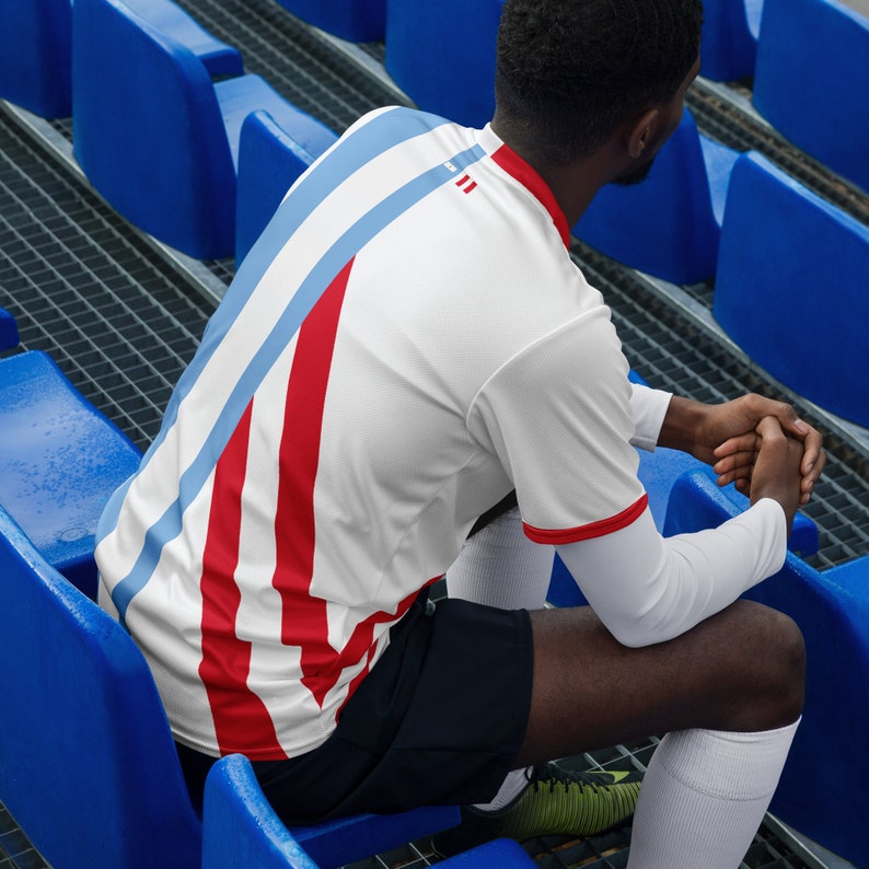 May include: A person wearing a white and red striped jersey with blue accents. The jersey has a white collar and short sleeves. The person is sitting on blue bleachers.