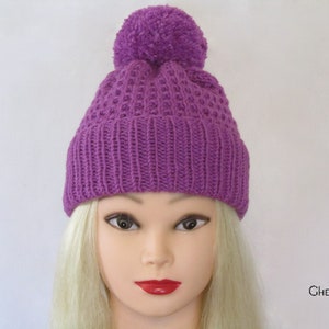 May include: Purple knitted beanie with a pom pom on top. The beanie has a textured pattern and is worn by a person with blonde hair.