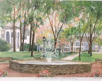 May include: A watercolor painting of a fountain in a park with green trees and white buildings in the background. The fountain is surrounded by a stone wall and there are benches on either side. The text "The Center Green - Glastonbury" is written in the bottom right corner of the painting.