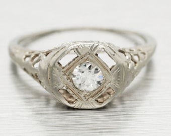 May include: A vintage diamond engagement ring with a delicate filigree design. The ring features a single round diamond set in a square-shaped bezel. The band is thin and has a delicate filigree design.