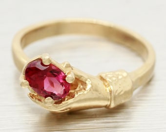 May include: A gold ring with a pink oval gemstone set in a hand design.