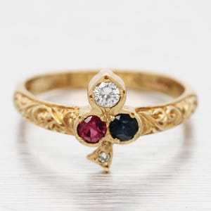 May include: A gold ring with an ornate band and a clover-shaped design. The clover features a central white diamond, a red gemstone, a blue gemstone, and a small diamond accent. The ring has an engraved floral pattern.