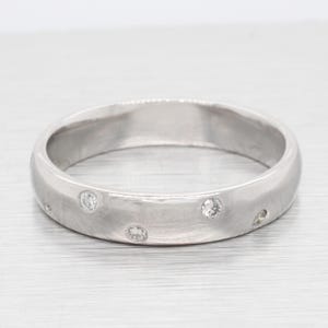 May include: A silver-colored ring with a brushed finish. The ring features several small, round, clear gemstones evenly spaced around the band. The ring is displayed on a light gray surface.