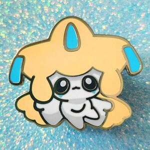 May include: A gold-colored enamel pin featuring a cartoon character with a light yellow head and body, white face, and blue accents. The character has large black eyes and a sad expression.