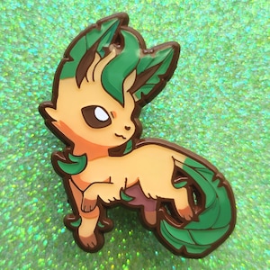 Puede incluir: Un colorido pin de esmalte con un personaje Leafeon de estilo caricatura. El pin tiene un cuerpo beige, detalles marrones y detalles de hojas verdes. El pin está sobre un fondo verde brillante.