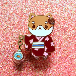 Puede incluir: Un pin de esmalte dorado con un perro Shiba Inu que lleva un kimono rojo y rosa con una faja azul. El perro sostiene una pequeña esfera de vidrio con un pez dorado dentro. El pin tiene una rosa y un amuleto colgante.
