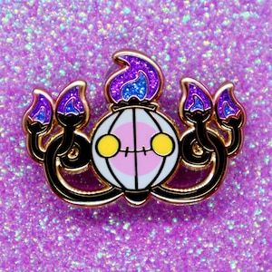 May include: Gold-colored enamel pin featuring a cartoon-style depiction of a purple and blue fire-type Pokemon with a round, white body and yellow eyes. The Pokemon has four purple and blue flames on its head and arms.