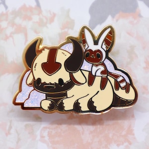 May include: A gold-tone enamel pin featuring a white and brown cartoon bison with a brown and white cartoon animal riding on its back. The bison has a brown marking on its forehead and the animal has a white face and brown ears.