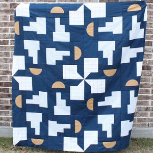 Op de afbeelding: Een marineblauwe quilt met een geometrisch patroon. Het ontwerp bevat witte vormen, sommige met gestreepte details, en bruine halve cirkel accenten. De quilt wordt getoond tegen een bakstenen muur.