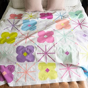 A colorful quilt with floral patterns and geometric shapes drapes over a bed with soft pillows. A leather bench sits at the foot, on a textured rug.