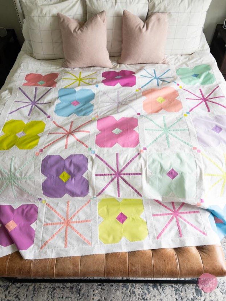 A colorful quilt with floral patterns and geometric shapes drapes over a bed with soft pillows. A leather bench sits at the foot, on a textured rug.