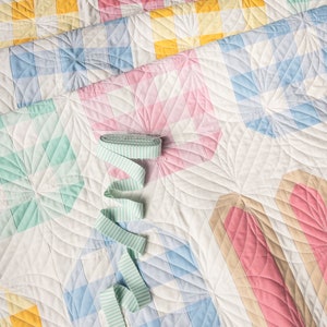 Colorful quilt with pastel squares, circles, and stripes. Featuring soft pinks, blues, yellows, and greens, a green striped ribbon lies on top.