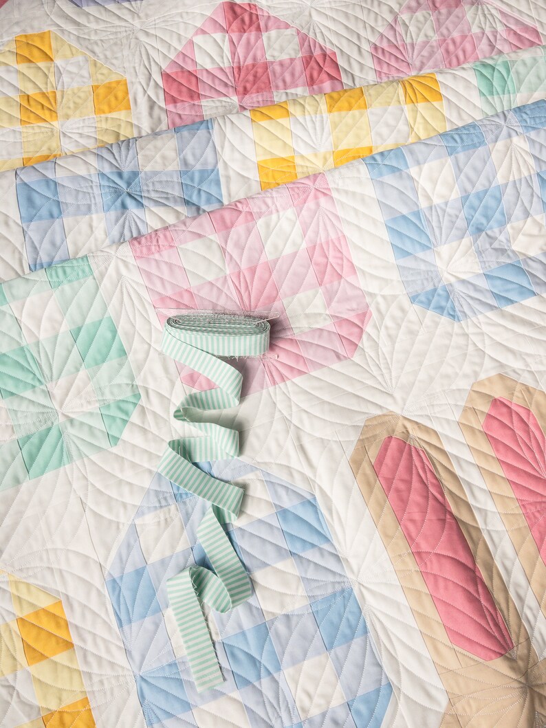 Colorful quilt with pastel squares, circles, and stripes. Featuring soft pinks, blues, yellows, and greens, a green striped ribbon lies on top.