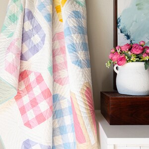 Puede incluir: Una colcha blanca con un dise&ntilde;o de patchwork que presenta varios patrones de cuadros vichy de colores pastel. La colcha est&aacute; doblada y drapeada sobre una escalera de madera.