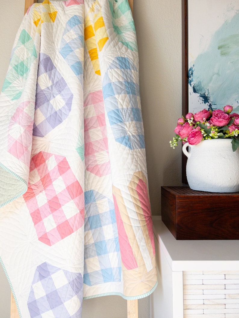 Puede incluir: Una colcha blanca con un dise&ntilde;o de patchwork que presenta varios patrones de cuadros vichy de colores pastel. La colcha est&aacute; doblada y drapeada sobre una escalera de madera.