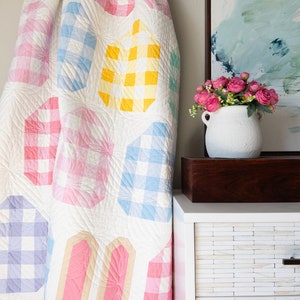 Quilt with pastel checkered patterns draped over a wooden ladder next to a white cabinet. A vase of pink flowers sits on the cabinet, below a soft abstract painting.