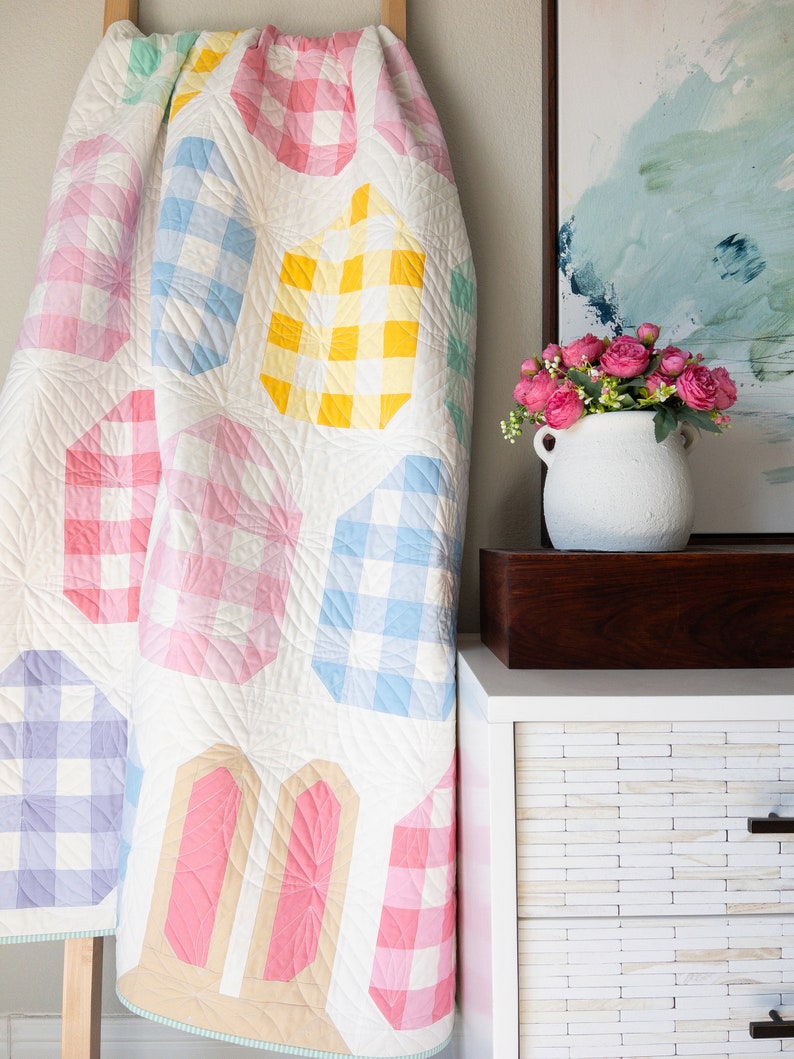 Quilt with pastel checkered patterns draped over a wooden ladder next to a white cabinet. A vase of pink flowers sits on the cabinet, below a soft abstract painting.