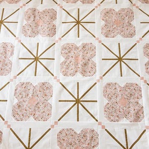 Quilt with alternating floral patterns and geometric designs. Features pink floral shapes and gold lines on white squares, creating a cozy, artistic feel.