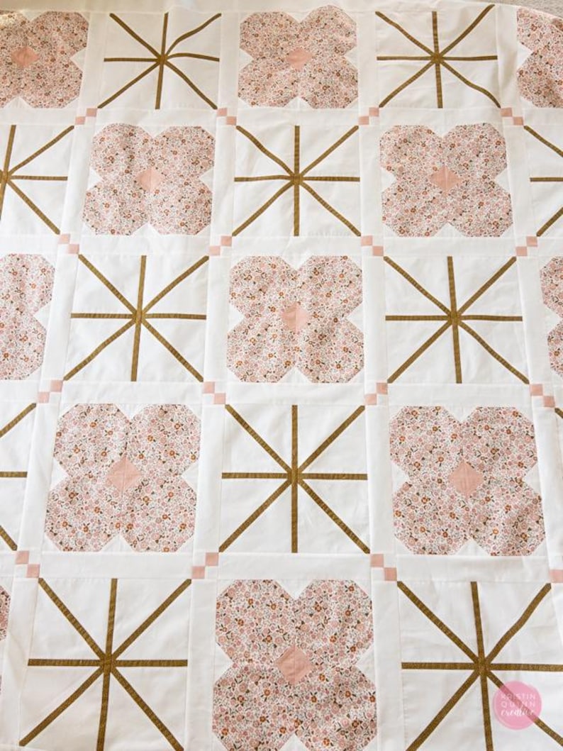 Quilt with alternating floral patterns and geometric designs. Features pink floral shapes and gold lines on white squares, creating a cozy, artistic feel.