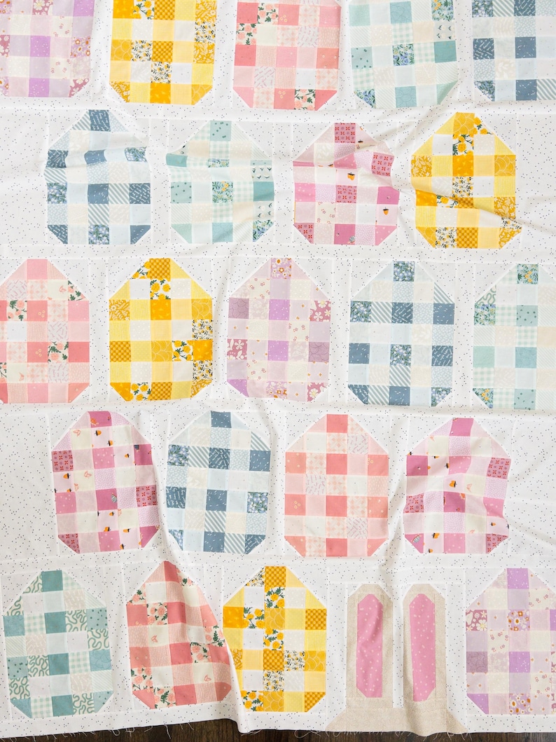 Colorful quilt with pastel checks in shades of pink, blue, yellow, and purple on a white speckled background. Cozy, homemade aesthetic.