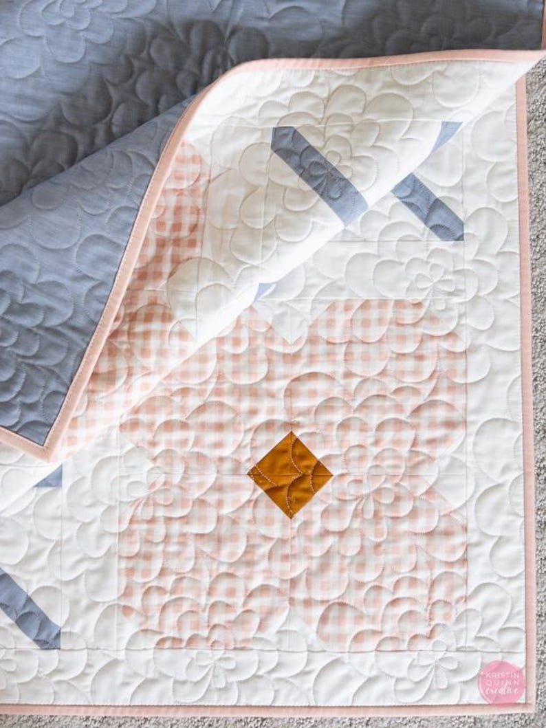 A quilt featuring a pink gingham pattern with blue floral stitching and a small mustard diamond accent. The quilt's soft tone is comforting and delicate.