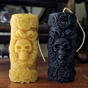 May include: Two decorative pillar candles, one black and one yellow, with a skull and floral design. The candles are made of beeswax and have a floral design with a skull in the center.