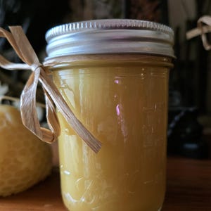 May include: A clear glass jar filled with a yellow liquid, topped with a silver lid. A natural raffia bow is tied around the jar. The jar is sitting on a wooden surface. A honeycomb-shaped candle is visible in the background.