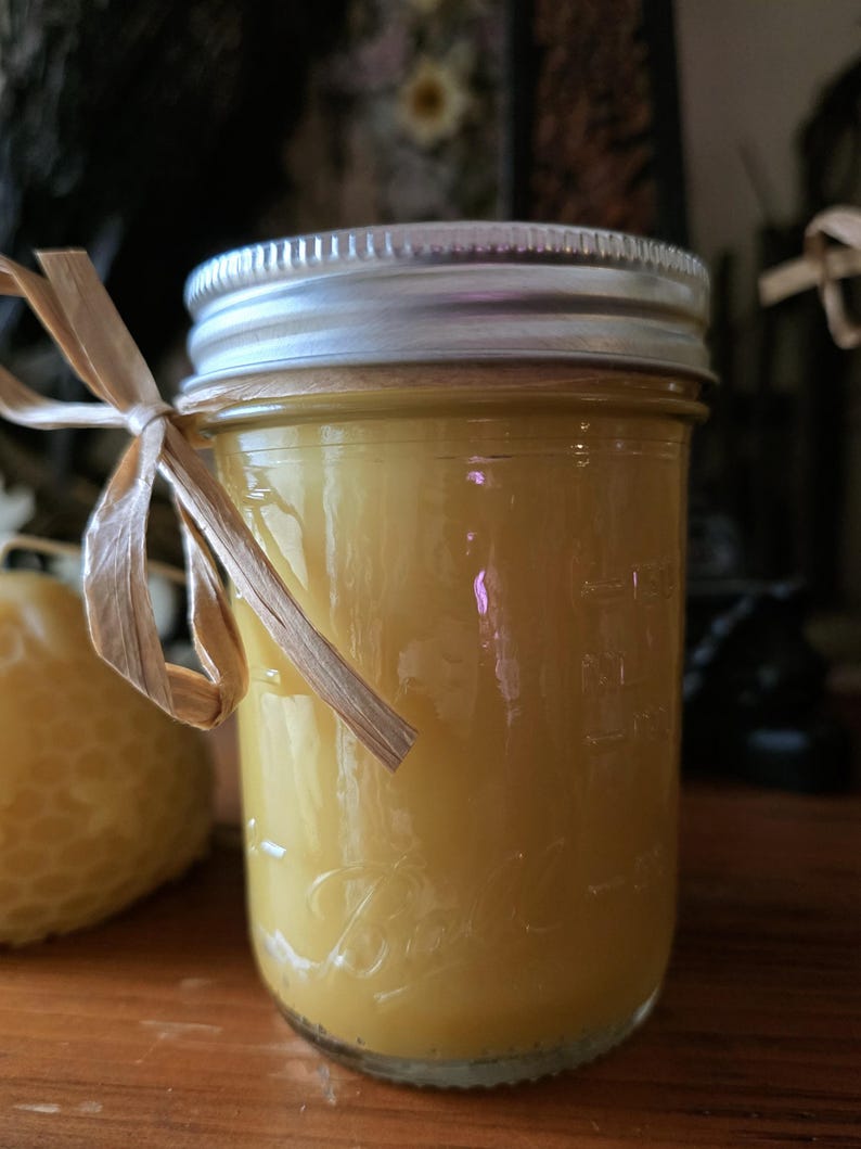 May include: A clear glass jar filled with a yellow liquid, topped with a silver lid. A natural raffia bow is tied around the jar. The jar is sitting on a wooden surface. A honeycomb-shaped candle is visible in the background.