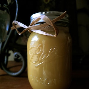 May include: A yellow-colored candle in a Ball Mason jar with a silver lid and a brown ribbon tied around the top. The jar is embossed with the Ball logo and the word "MASON". The candle is on a wooden surface.