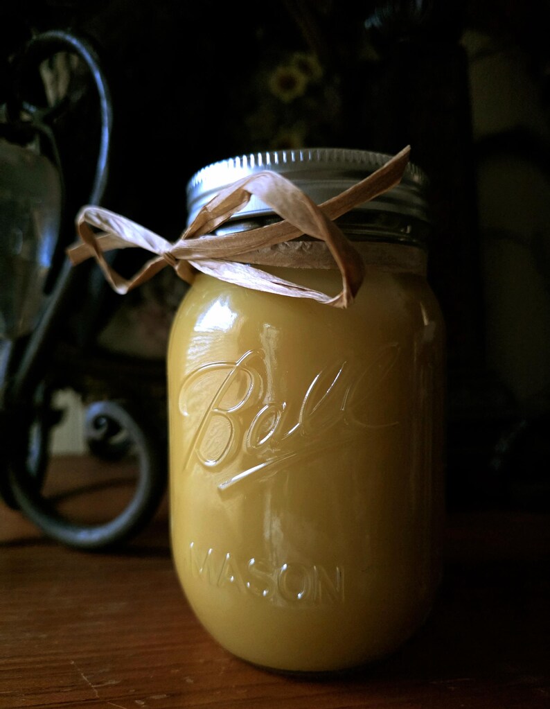May include: A yellow-colored candle in a Ball Mason jar with a silver lid and a brown ribbon tied around the top. The jar is embossed with the Ball logo and the word "MASON". The candle is on a wooden surface.