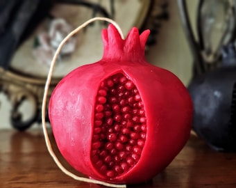 May include: A vibrant red pomegranate-shaped candle with a detailed, open design revealing the seed-filled interior. A cream-colored string is attached. The candle is on a wooden surface, with a blurred background featuring a decorative element.