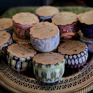 May include: A collection of decorative tin candles with patterned designs in various colors, including blue, red, and green. Each candle has a paper label on top. The candles are arranged on a silver tray.