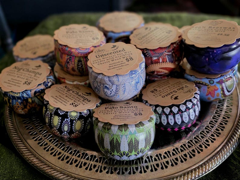 May include: A collection of decorative tin candles with patterned designs in various colors, including blue, red, and green. Each candle has a paper label on top. The candles are arranged on a silver tray.