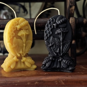 May include: Two oval-shaped beeswax candles with a bee and flower design. One candle is yellow and the other is black.