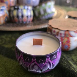 May include: A close-up of a scented candle in a decorative tin. The candle has a wooden wick and is surrounded by a purple and black patterned design. Other candles with colorful designs are in the background, suggesting a collection of candles.